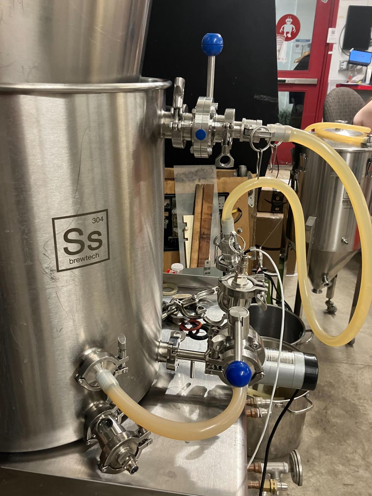 This close-up shot shows a large, cylindrical stainless steel brewing vessel from "Ss Brewtech" with various tubes and valves attached. A pale yellow hose extends from a valve near the top right, looping down and connecting to another valve assembly near the bottom of the vessel.