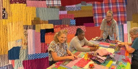 Four people sit around a table hand-stitching colorful fabric pieces, with a large patchwork quilt shown behind them.