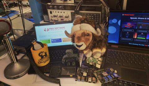 A desk cluttered with laptops showing the Free Software Foundation website, a GNU plush, a Tux penguin figurine, circuit boards, and vintage radio equipment.