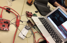 A table is covered with electronics gear including a laptop running code, red circuit boards, a white breadboard, and a tangle of jumper wires connecting components. Two people are working nearby, surrounded by laptops and tools, suggesting an active hardware tinkering or coding session.