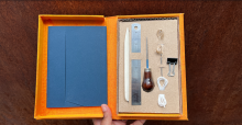 On a red background, an open clam shell box containing a set of book-making tools, nested into a cardboard frame