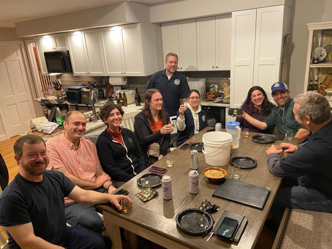 A group of smiling friends sit around a table holding and toasting with glasses of beer.