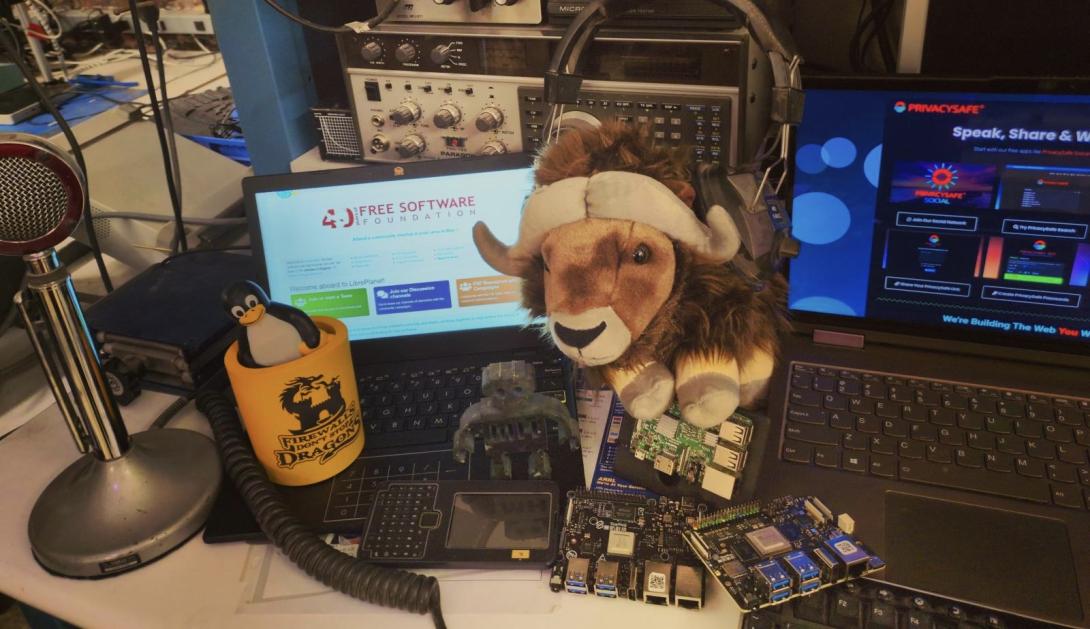 A desk cluttered with laptops showing the Free Software Foundation website, a GNU plush, a Tux penguin figurine, circuit boards, and vintage radio equipment.