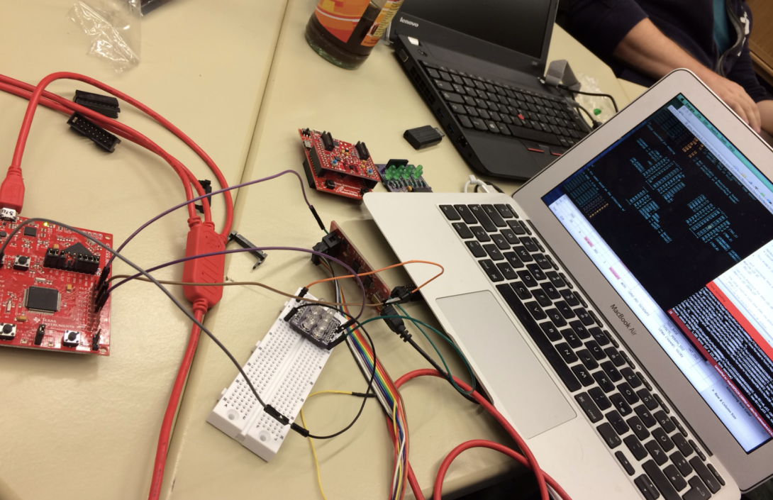 A table is covered with electronics gear including a laptop running code, red circuit boards, a white breadboard, and a tangle of jumper wires connecting components. Two people are working nearby, surrounded by laptops and tools, suggesting an active hardware tinkering or coding session.
