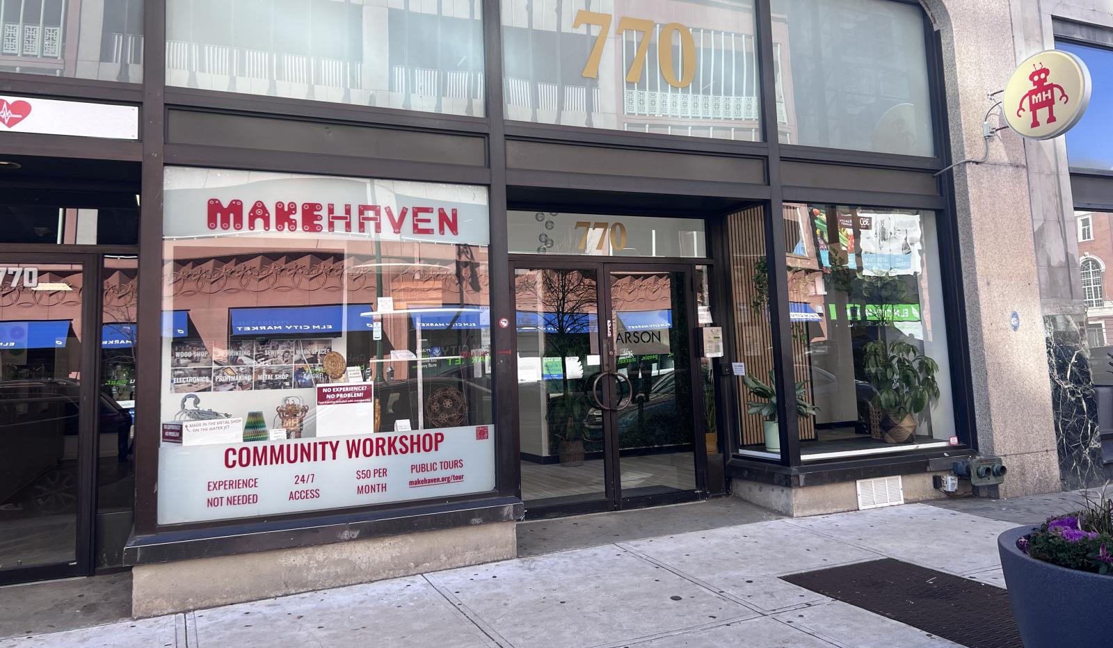 The main entrance of MakeHaven is seen at 770 Chapel Street. A large gold 770 is above the doorway. To the right of the door is a storefront window filled with projects made at MakeHaven including a cutting board, a ceramic mug, a metal scorpion, a gold cast Harry Potter resin piece and more.