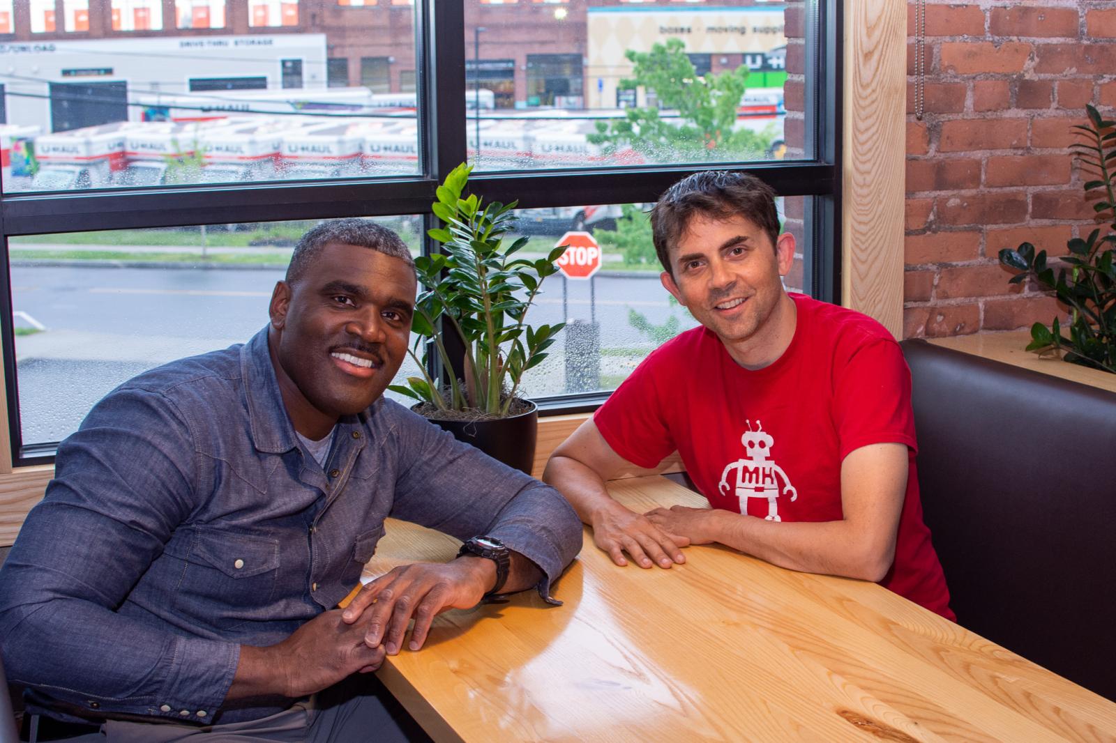 Two people sitting at a table, smiling. On the left is Brent Peterkin, a black man wearing a blue shirt; on the right is J.R. Logan, a white man wearing a red shirt with a MakeHaven robot on it.
