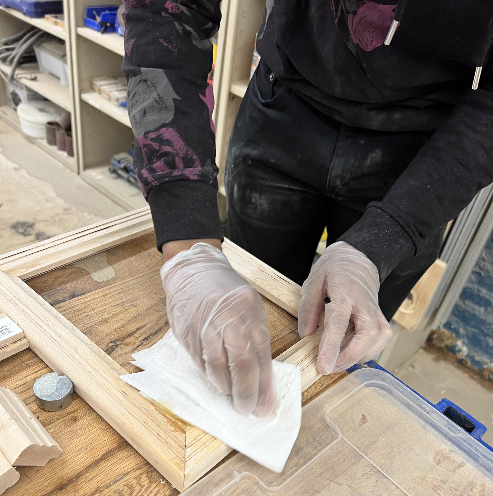 The hands of a black person wearing gloves add a finishing varnish to a wooden frame they just made on a workbench.